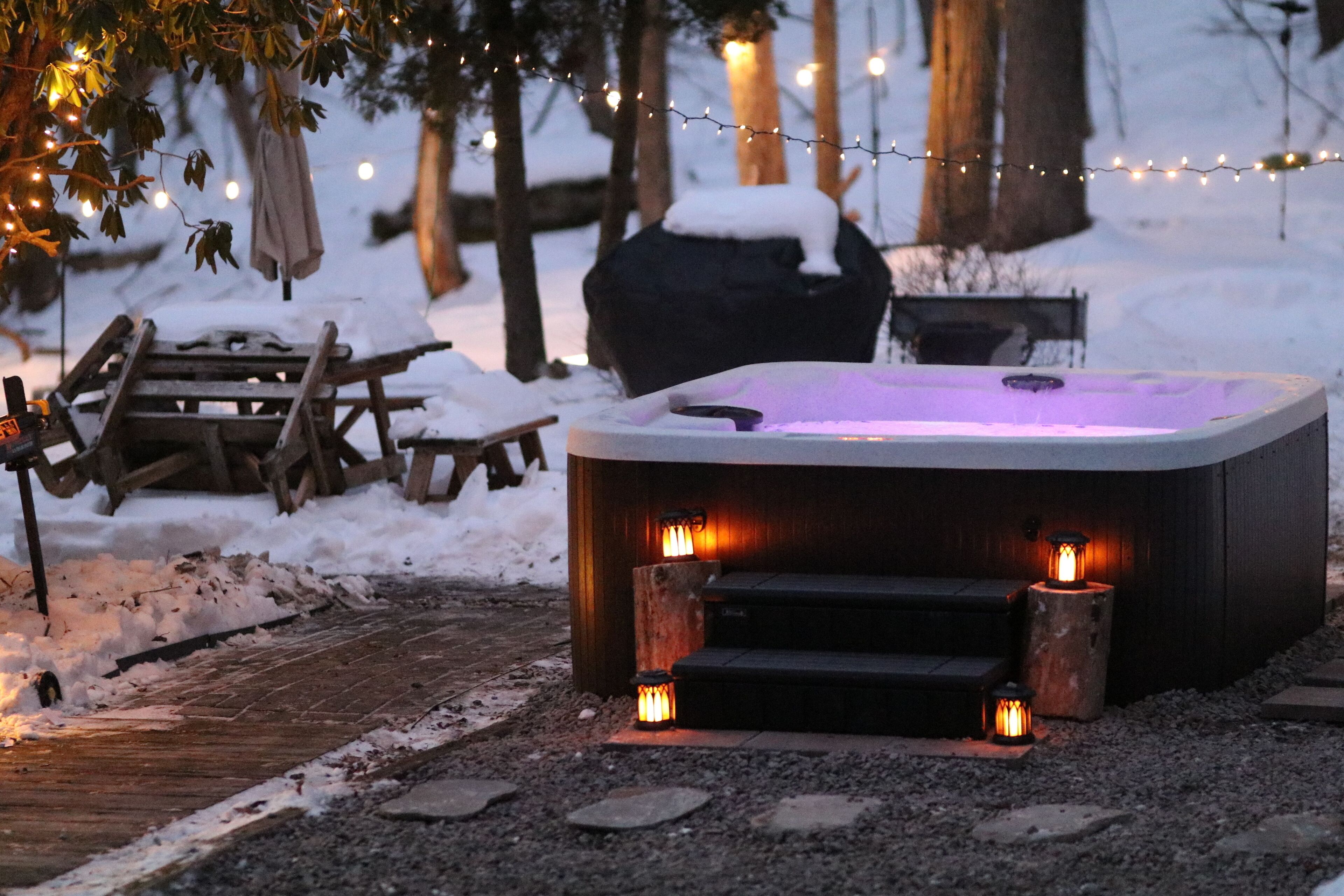 A hot tub with steps and lanterns, surrounded by snow and trees.