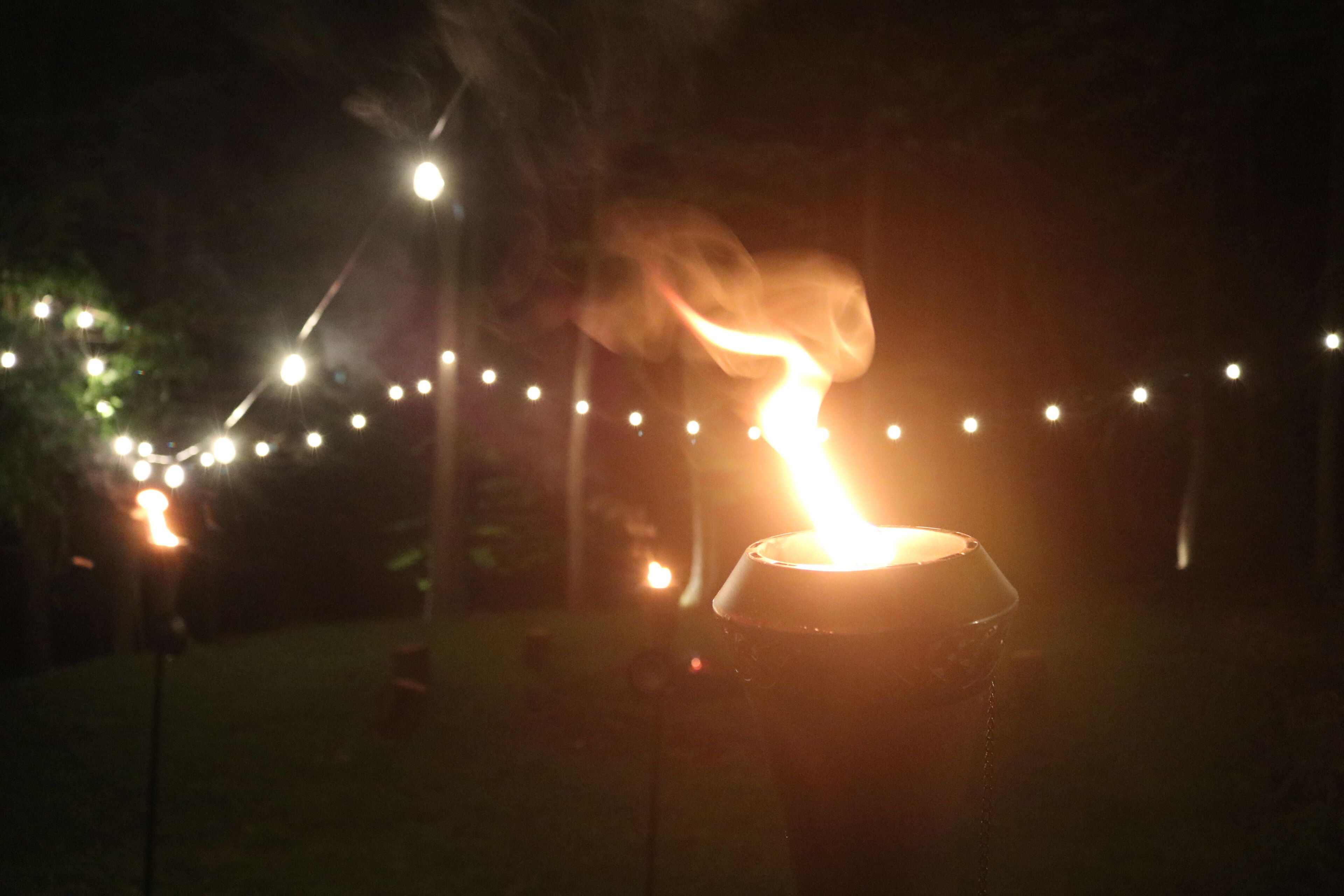 A candle in a glass holder outdoors at night with string lights in the background.