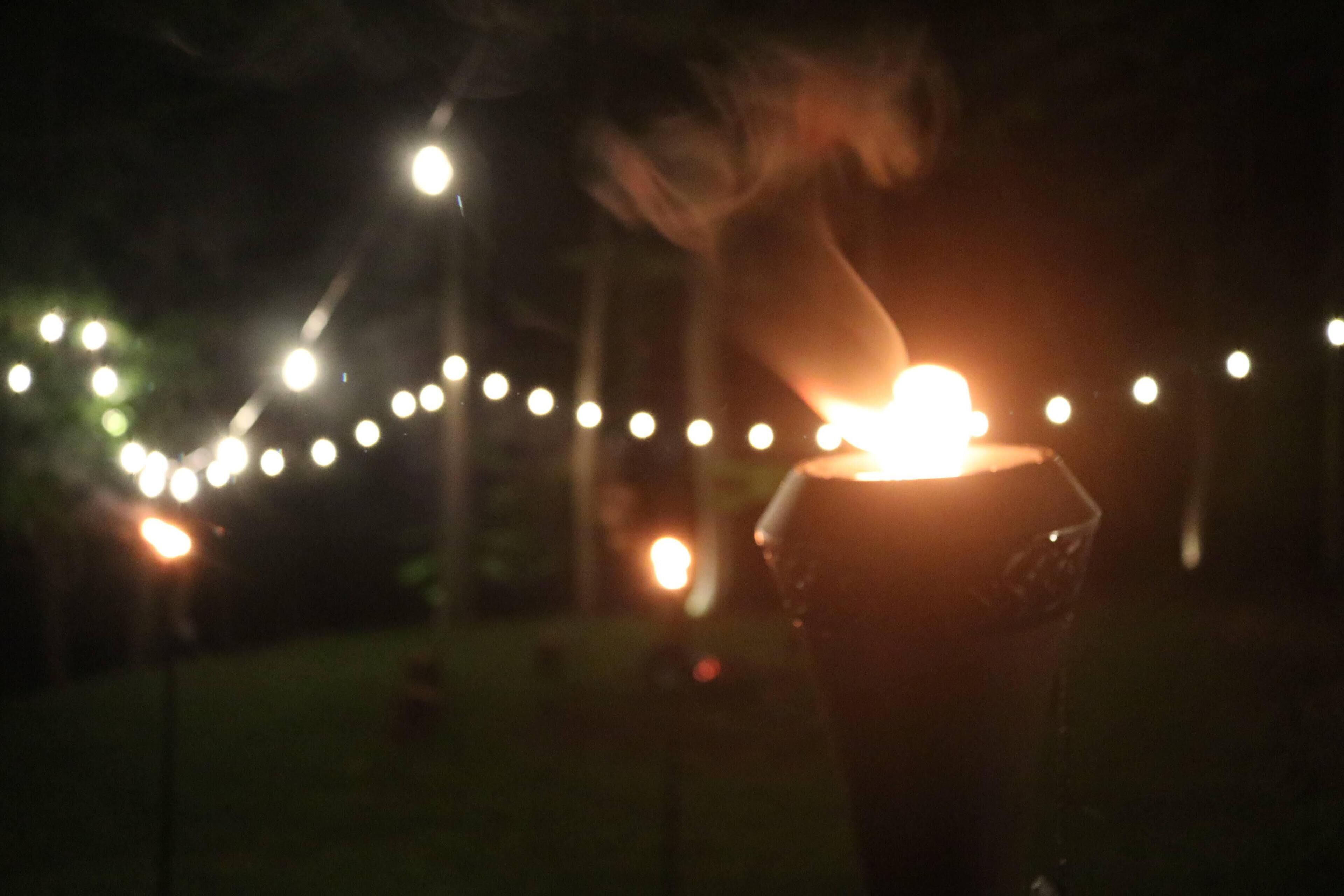 Un espace extérieur faiblement éclairé, décoré de guirlandes lumineuses, où l’on aperçoit une personne tenant un objet lumineux.