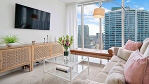 A modern living room with a large window, a flat-screen TV, a wooden cabinet, a glass coffee table, and a sofa with cushions. - Open Plan Brickell Suite with Workspace (Miami)