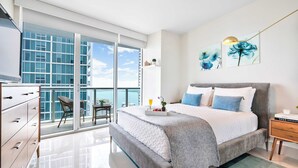 A modern bedroom with a large bed, a nightstand, a dresser, and a balcony with a view of the city. - Open Plan Brickell Suite with Workspace (Miami)