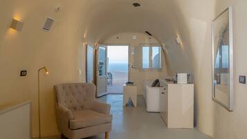 Amplia habitación con un techo alto abovedado, un sillón capitonado, un escritorio con una silla y vista al mar a través de un gran ventanal.
