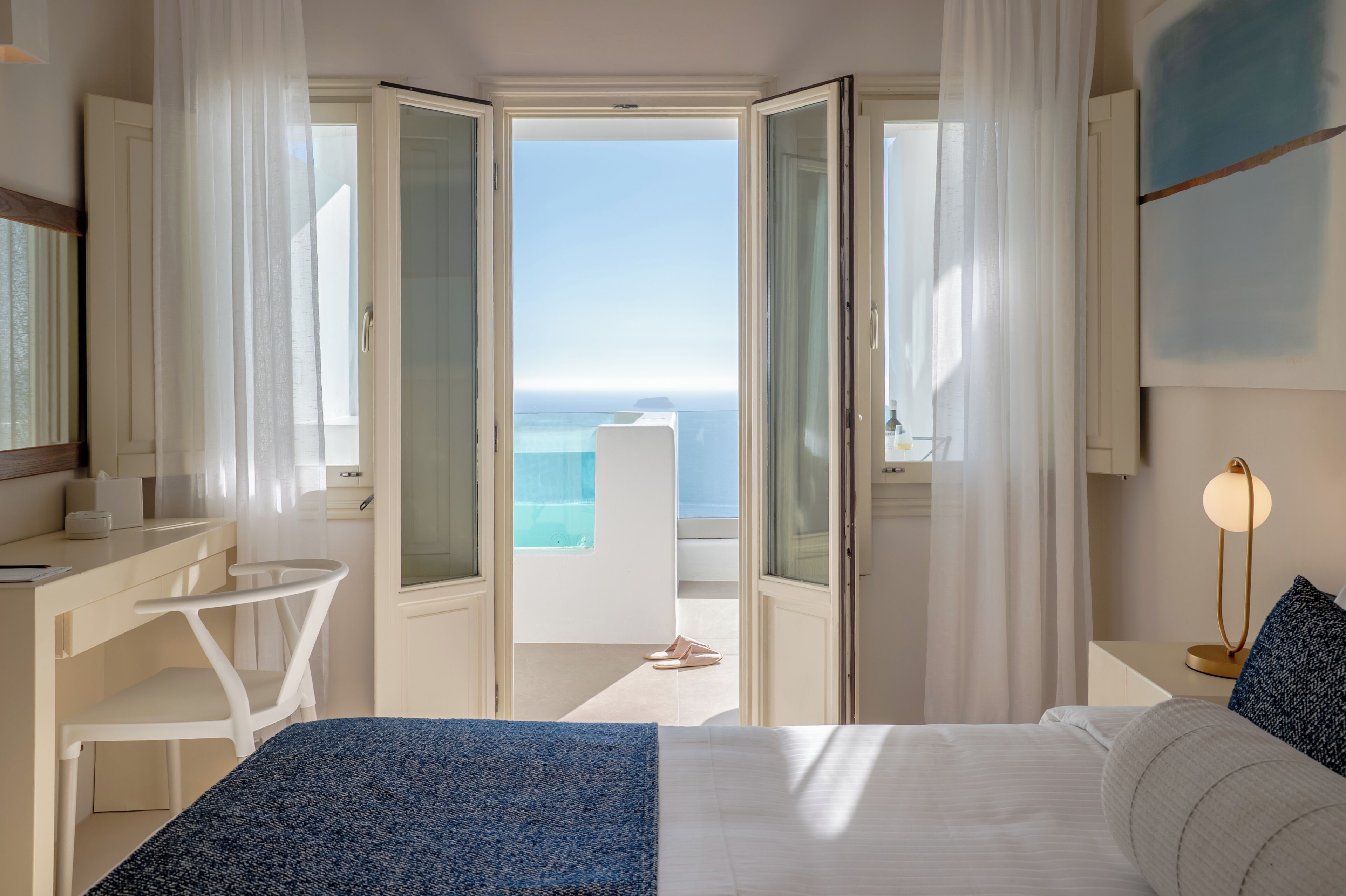 A hotel room with a bed, a desk, a chair, a bedside table, and a view of the ocean.