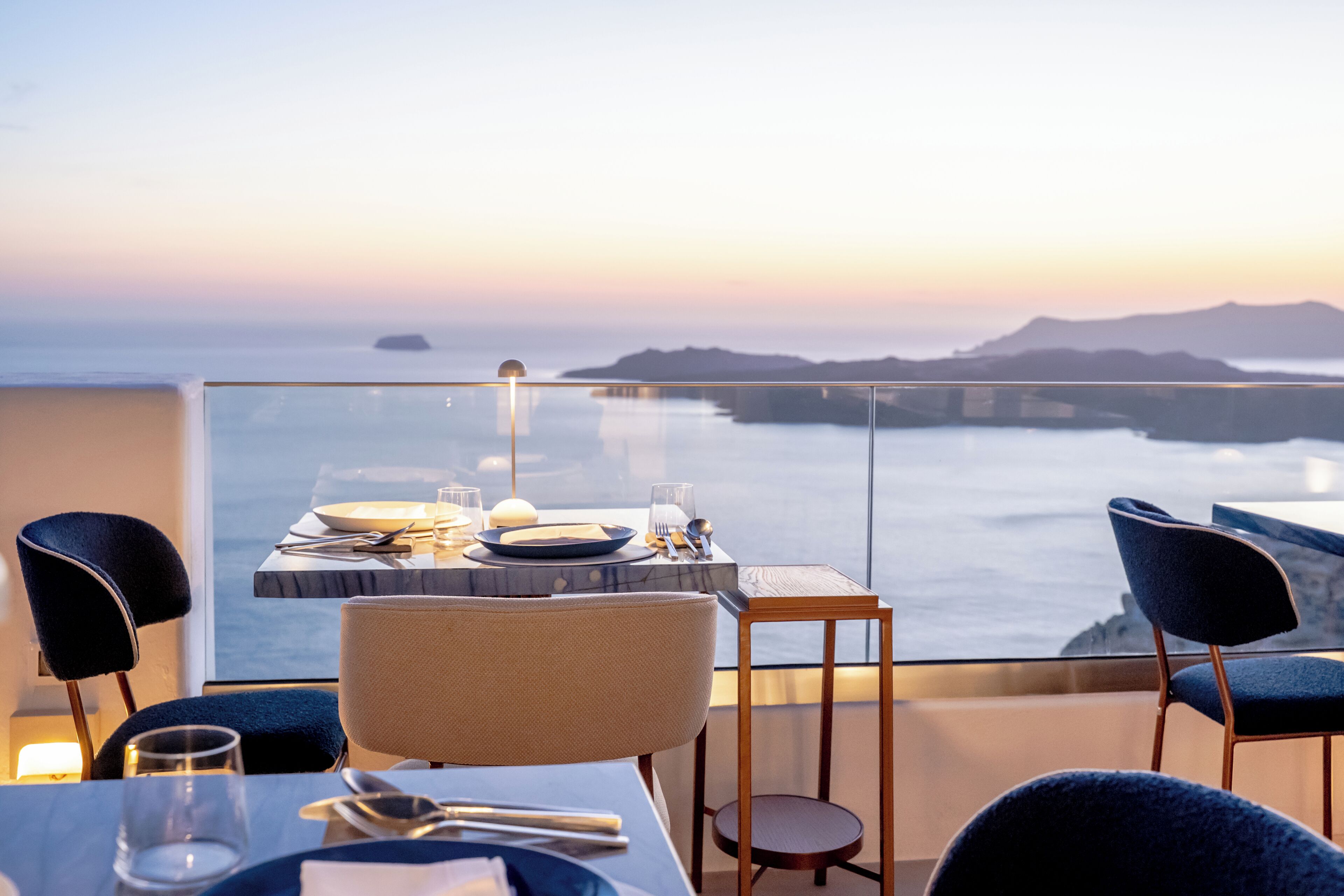 Mesa lista con platos, cubiertos y vasos, con vistas a la costa, cielo despejado y tierra distante.