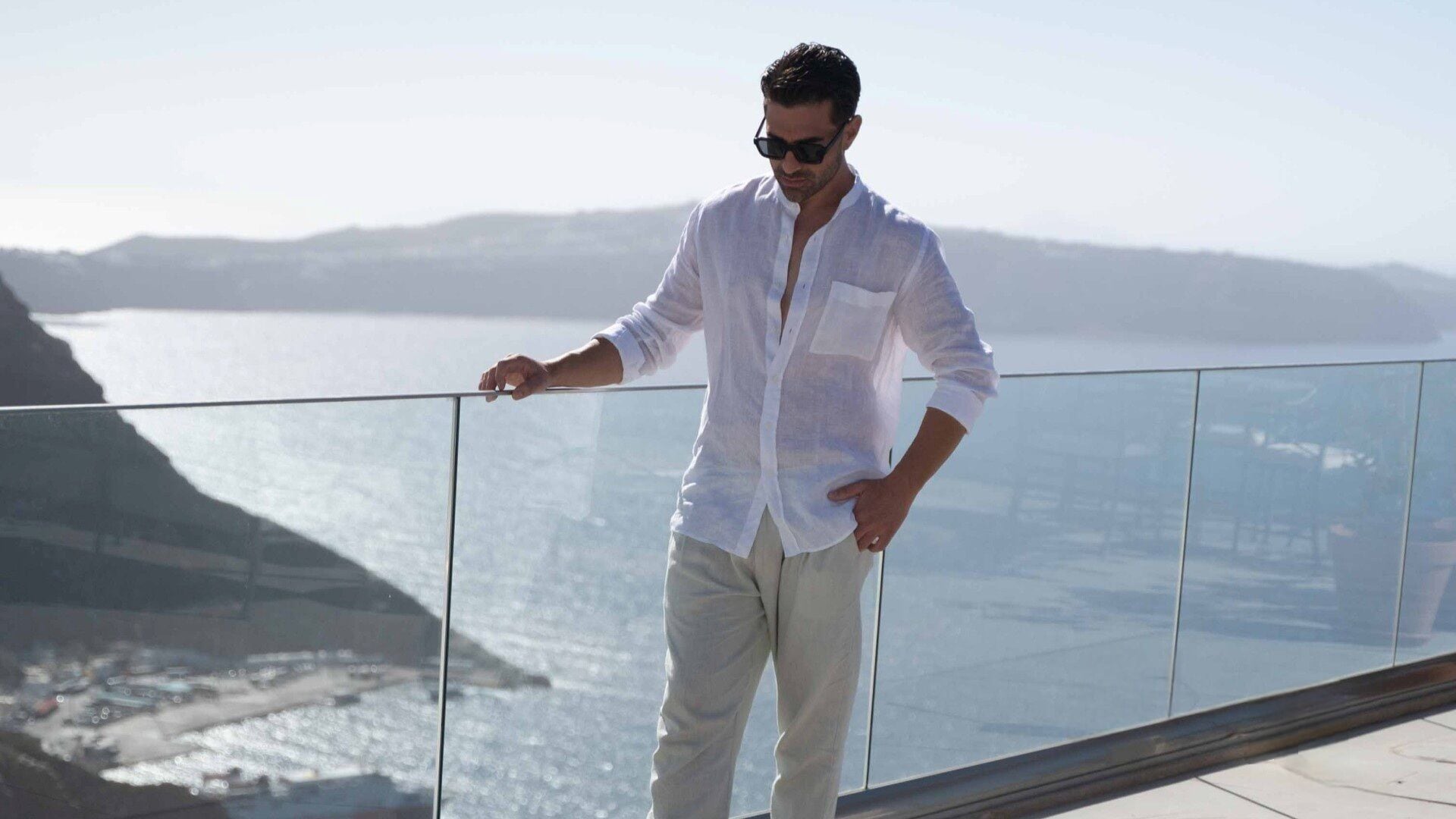 Um homem está em uma varanda, com vista para a paisagem costeira, o mar e colinas ao longe.