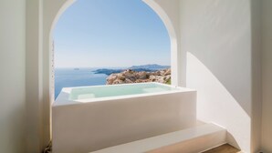 A white bathtub with a clear view of the sea and rocky landscape through an arched window. - Apeiron Blue by Apeiron Hotels (Santorini)