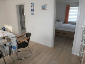A room with a bed, a desk with a chair, a small table with a plant, and a window with curtains.