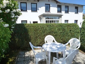 Ein weißes Gebäude mit vielen Fenstern, einem Heckenstreifen und einer Terrasse mit weißen Plastikstühlen und einem Tisch.