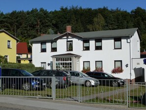 A two-story building with a central entrance, multiple windows, and a parking area with cars.