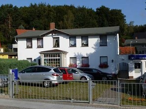 Ein zweistöckiges Gebäude mit weißer Fassade, einem zentralen Eingang mit Tür und Fenstern sowie einer kleinen Veranda.