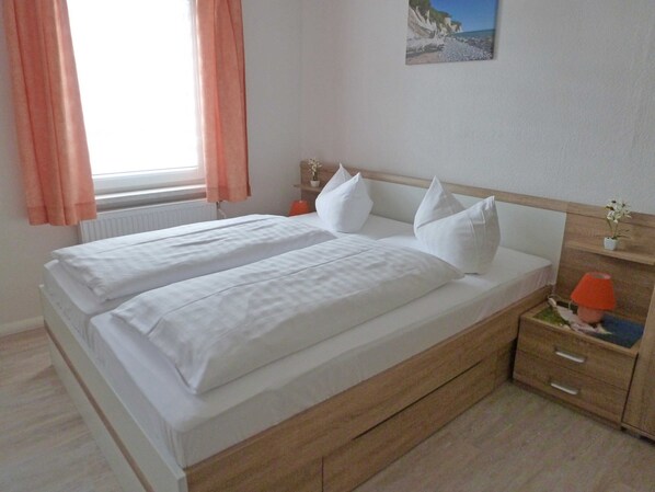 A double bed with white linens, a wooden headboard, a bedside table with a lamp, and a window with curtains.