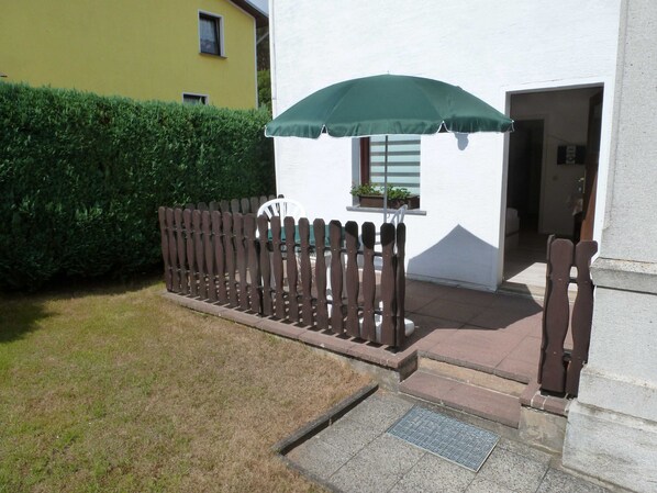 Ein Holzzaun, ein grüner Sonnenschirm, ein weißes Gebäude und ein Hecken.