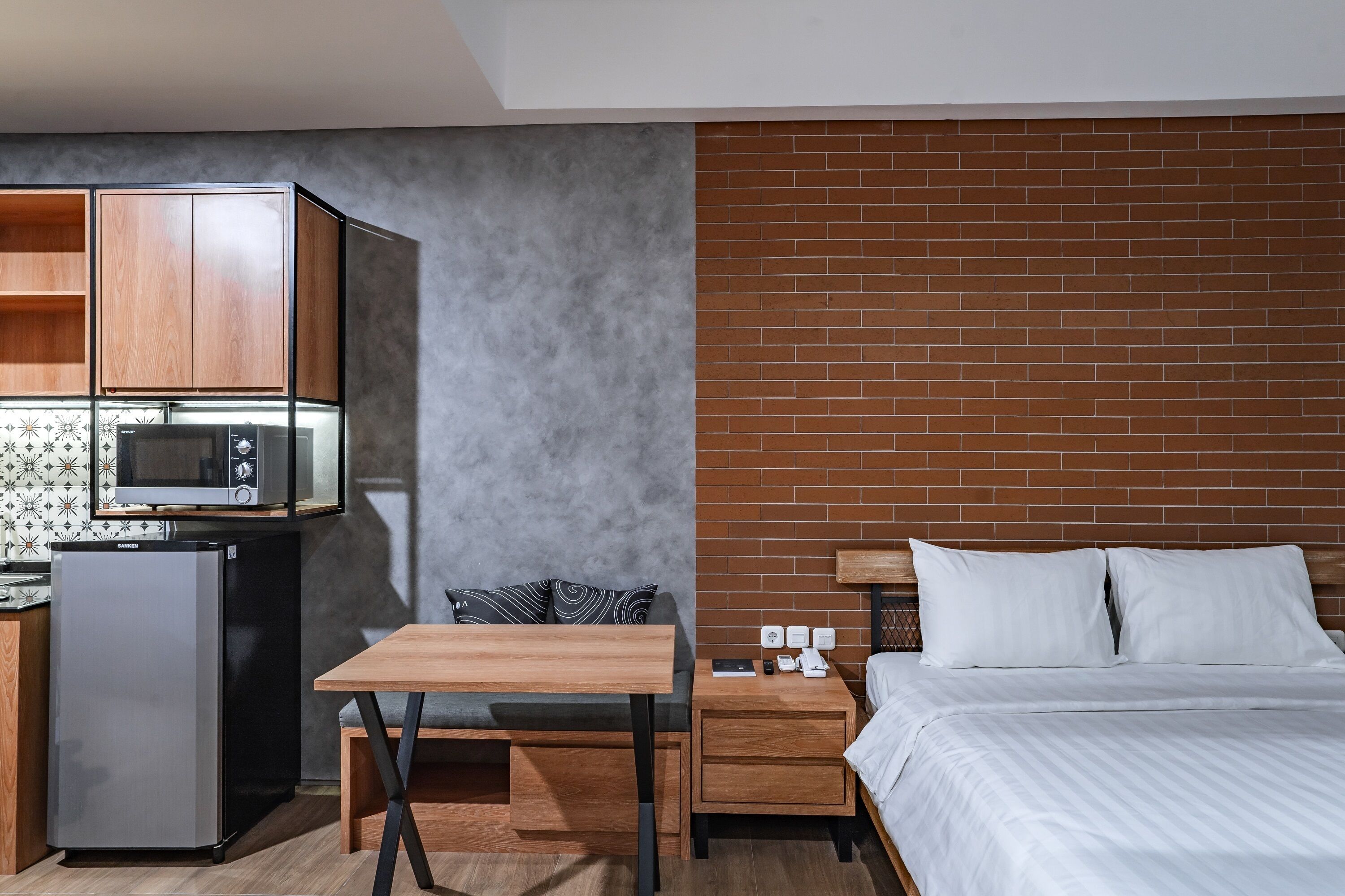 A compact hotel room with a kitchenette, a bed with white bedding, a small table, and a brick accent wall.