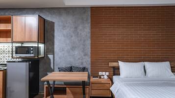 A compact hotel room with a kitchenette, a bed with white bedding, a small table, and a brick accent wall.