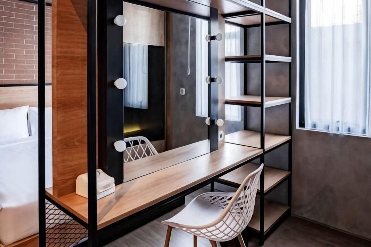 A room with a mirror, vanity, and shelves; a chair; a bed; and a window with curtains.