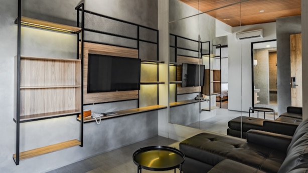 A modern living room with a large flat-screen TV mounted on a wooden shelf, a black leather sofa, and a small round coffee table. - LOA Living Solo Baru (Grogol)