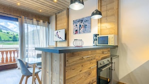 A modern kitchen with wooden cabinetry, a built-in microwave, and a stainless steel oven. There is a dining area with a glass table and chairs, and a balcony with a view of the countryside. - Large Studio Cabin, Balcony, Parking, La Clusaz Center (La Clusaz)