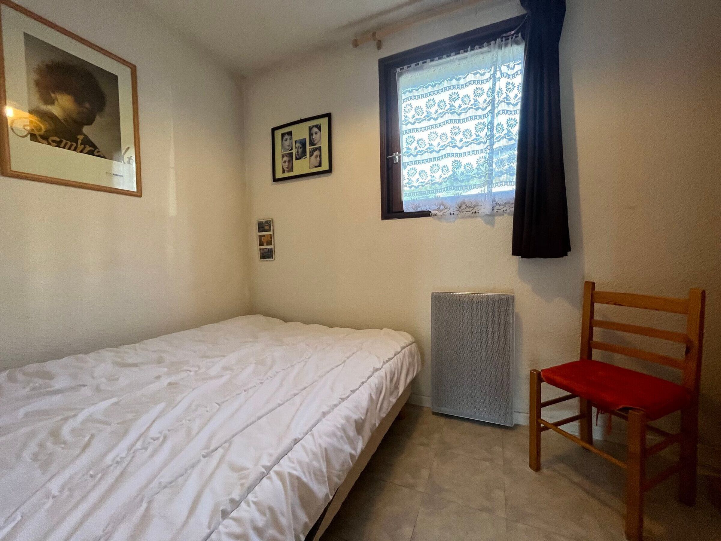 A small, single-bed room with a radiator, a wooden chair with a red cushion, and a window with patterned curtains.