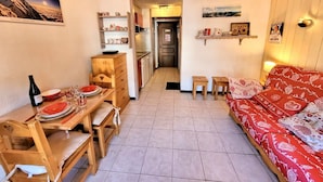 A cozy living room with a red patterned sofa, a wooden dining table with chairs, and a kitchen in the background. - Central studio, near slopes, ideal for 2 (Les Gets)