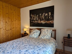 Un dormitorio con cama, mesita de noche, lámpara, techo de madera y una pintura de un paisaje urbano en la pared.
