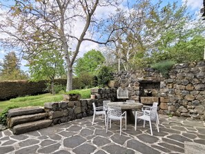 Un patio al aire libre con suelo de piedra, sillas de metal blanco, una mesa redonda y un muro de piedra con una jaula de pájaros.