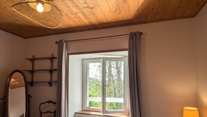 A bedroom with a bed, a window with curtains, a bedside table with a lamp, a chair, and a mirror.