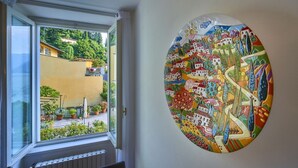 A room with a large window overlooking a garden, a colorful abstract painting on the wall, and a radiator under the window. - Casa Lido beautiful lake view (Varenna)