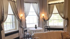 A cozy room with a sofa, a table with a chair, and two windows with curtains. - Book all 4 units in this National Historic Landmark in the Village of Geneseo (Geneseo)