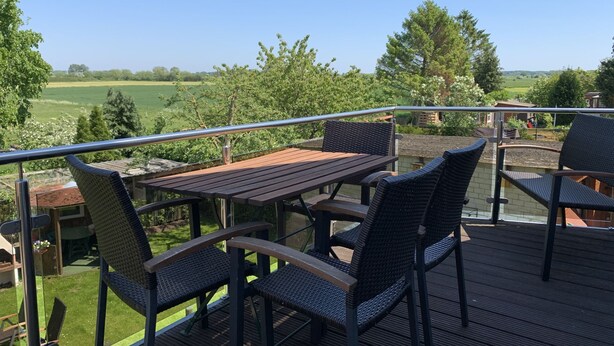 Una terraza al aire libre con mesa de madera y sillas de mimbre, con vista a un paisaje verde.