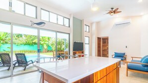Una cocina moderna con una isla grande, un comedor con sillas, un televisor y vistas a una piscina y follaje exterior.