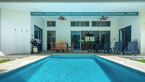 Una piscina con sillones de descanso y una mesa, rodeada por un edificio con ventanas amplias y un ventilador de techo.