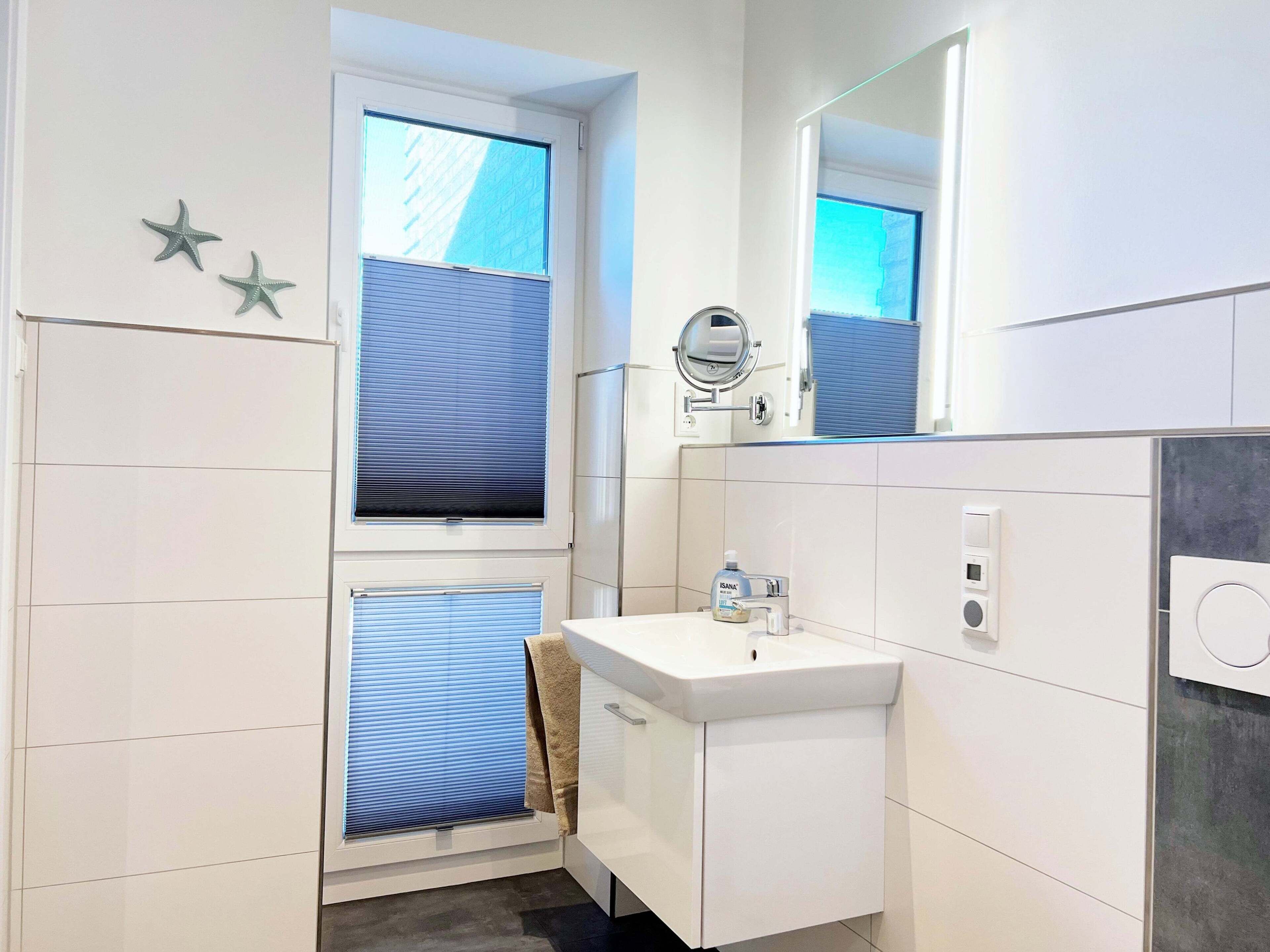 A modern bathroom with a white sink, a mirror, and a window with blinds.