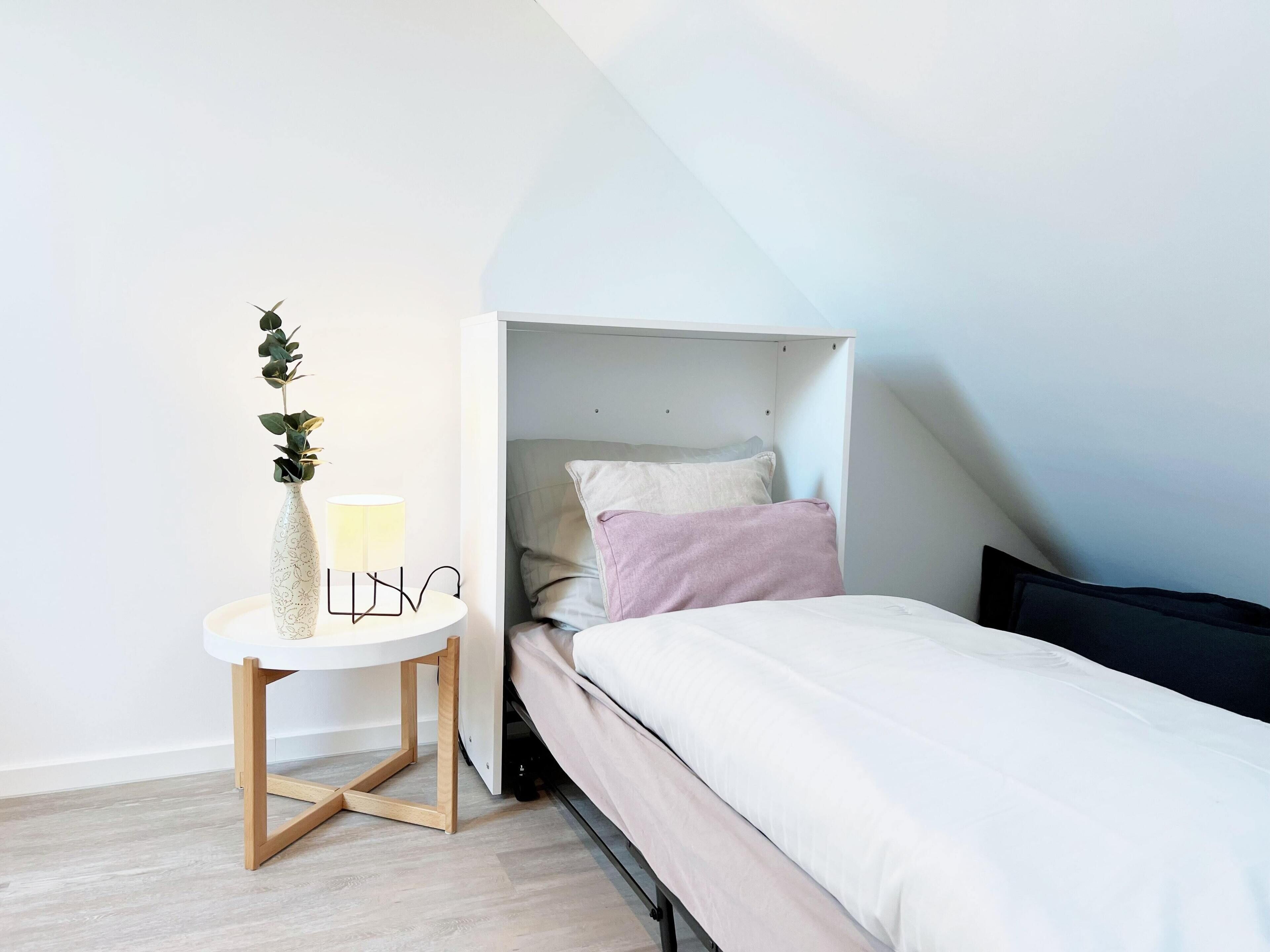 A small, modern bedroom with a bed, a bedside table with a lamp, and a potted plant.