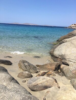 Une plage rocheuse bordée d’une eau d’un bleu limpide et dominée par des collines lointaines.
