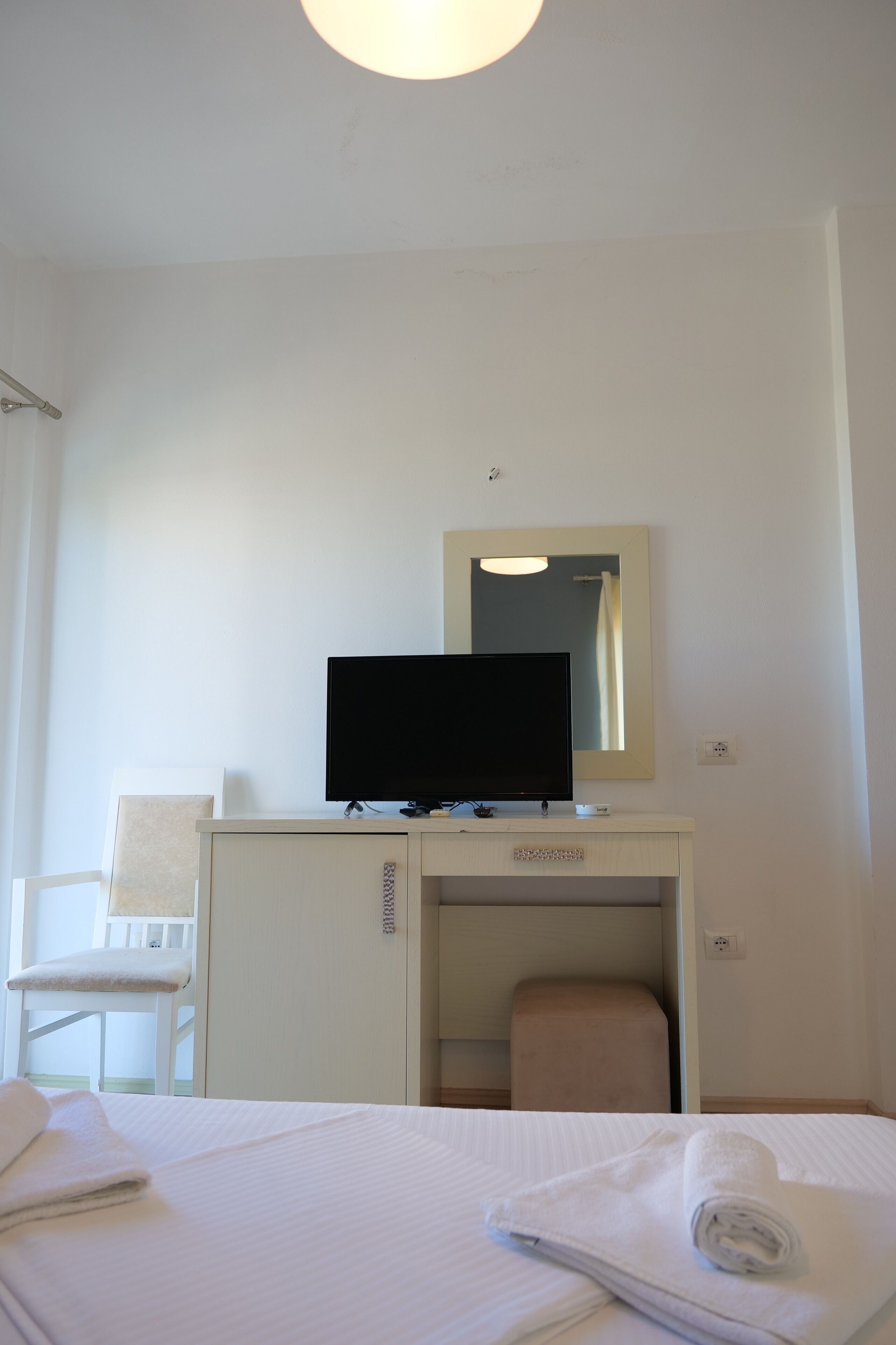 A hotel room with a bed, a desk with a television, a chair, and a small stool.