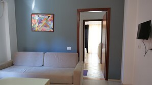 A living room with a beige sofa, a small table, a colorful abstract painting, and a TV mounted on the wall. - Privé City Hotel (Sarandë)