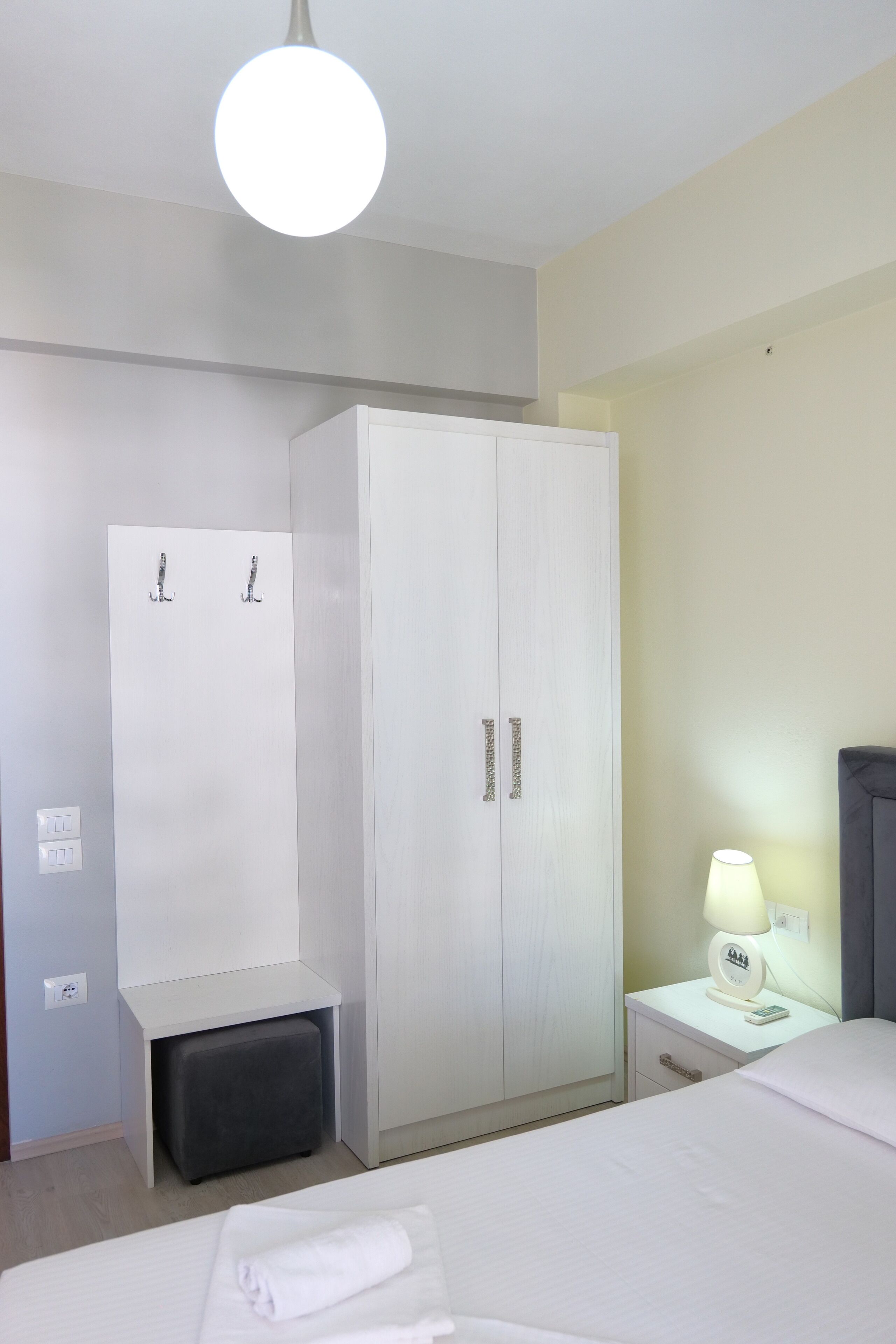A hotel room with a bed, a wardrobe, a nightstand with a lamp, and a small bench.