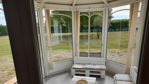 A conservatory with a bench and a small table, offering a view of a garden. - Maison Toute Équipée Avec Piscine et Pool House (Gaillan-en-Médoc)