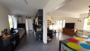A spacious living area with a kitchen, dining table with colorful placemats, a television, and a large window. - Maison Toute Équipée Avec Piscine et Pool House (Gaillan-en-Médoc)
