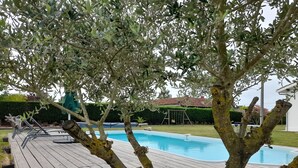 A wooden deck with a swimming pool, surrounded by olive trees and a well-maintained lawn. - Maison Toute Équipée Avec Piscine et Pool House (Gaillan-en-Médoc)