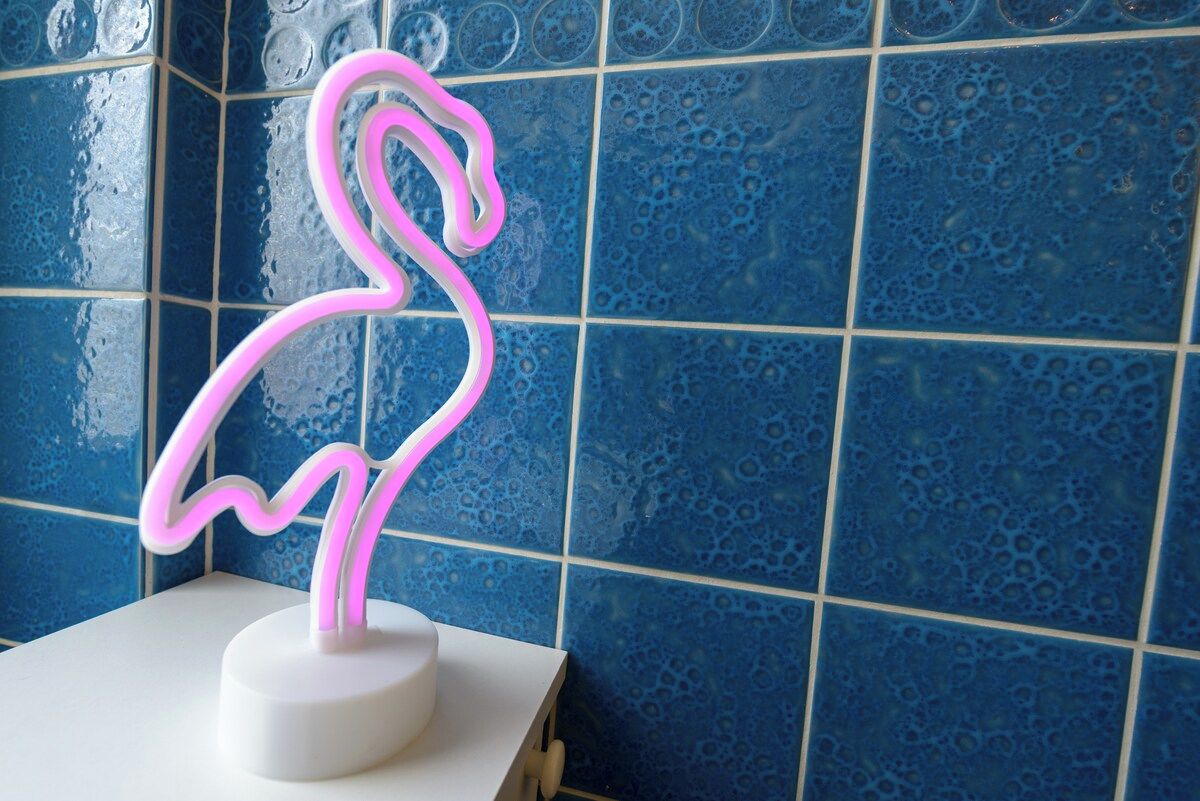 Een neon vlamingo bord op een witte basis tegen een blauwe betegelde muur.