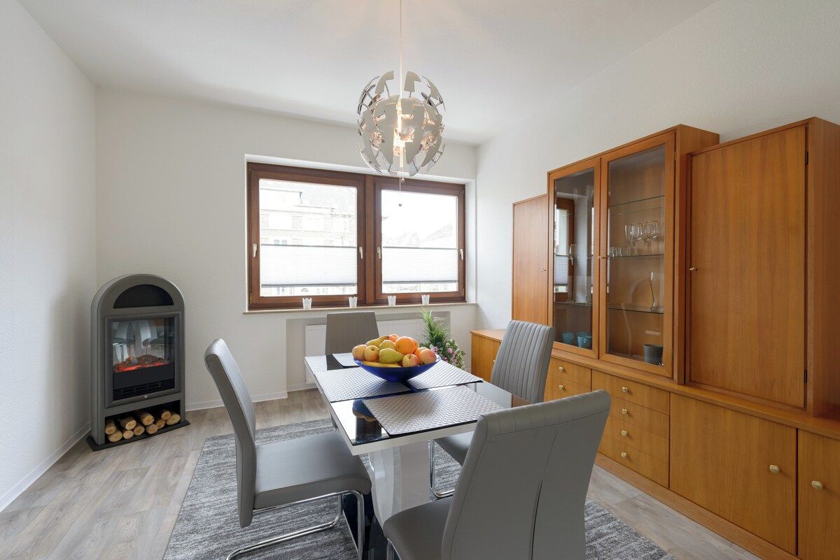 A dining area with a table, chairs, a bowl of fruit, a wood-burning stove, and a cabinet with glass doors.