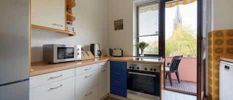 A compact kitchen with a microwave, toaster, and stove, a small dining area with a chair, and a view of a building through a window.
