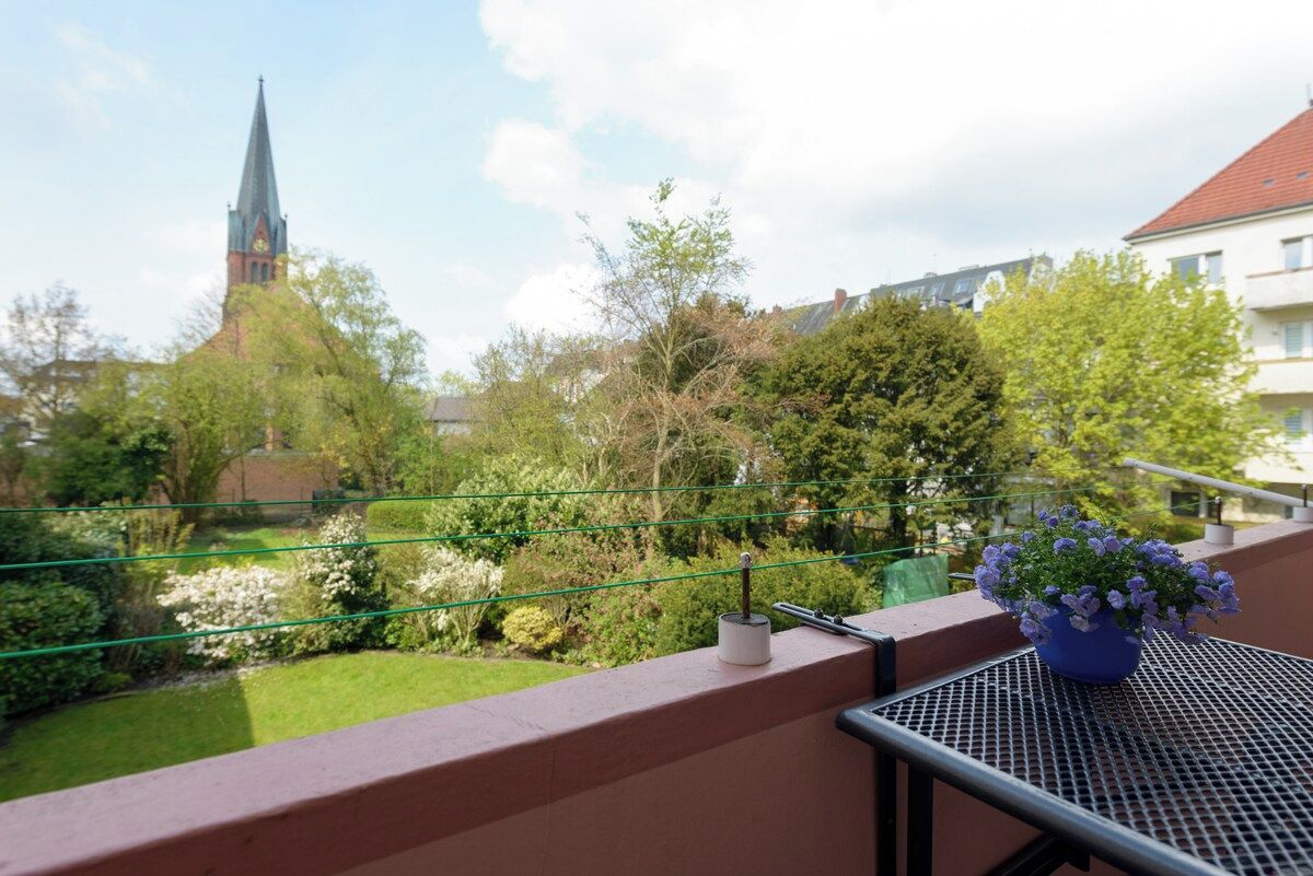 A balcony with a view of a church spire, greenery, and a potted plant.