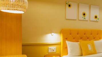 Une chambre d’hôtel avec une tête de lit jaune à motifs capitonnés, un lit avec une literie blanche, un luminaire suspendu lumineux et trois œuvres d’art murales représentant des tournesols.