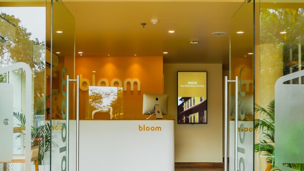 A reception area with a glass partition, a reception desk, and a digital screen displaying 'Kochi The Waterfront Gallery'. - Bloom Hotel - Worli (Mumbai)