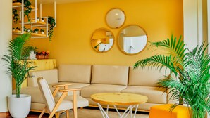 A modern living room with a beige sofa, a round wooden coffee table, and a white armchair. There are three wall-mounted mirrors and a variety of potted plants. - Bloom Hotel - Worli (Mumbai)