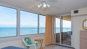 Una stanza con un letto, un tavolino, una sedia, una ventola a soffitto e vista sull'oceano attraverso ampie finestre.