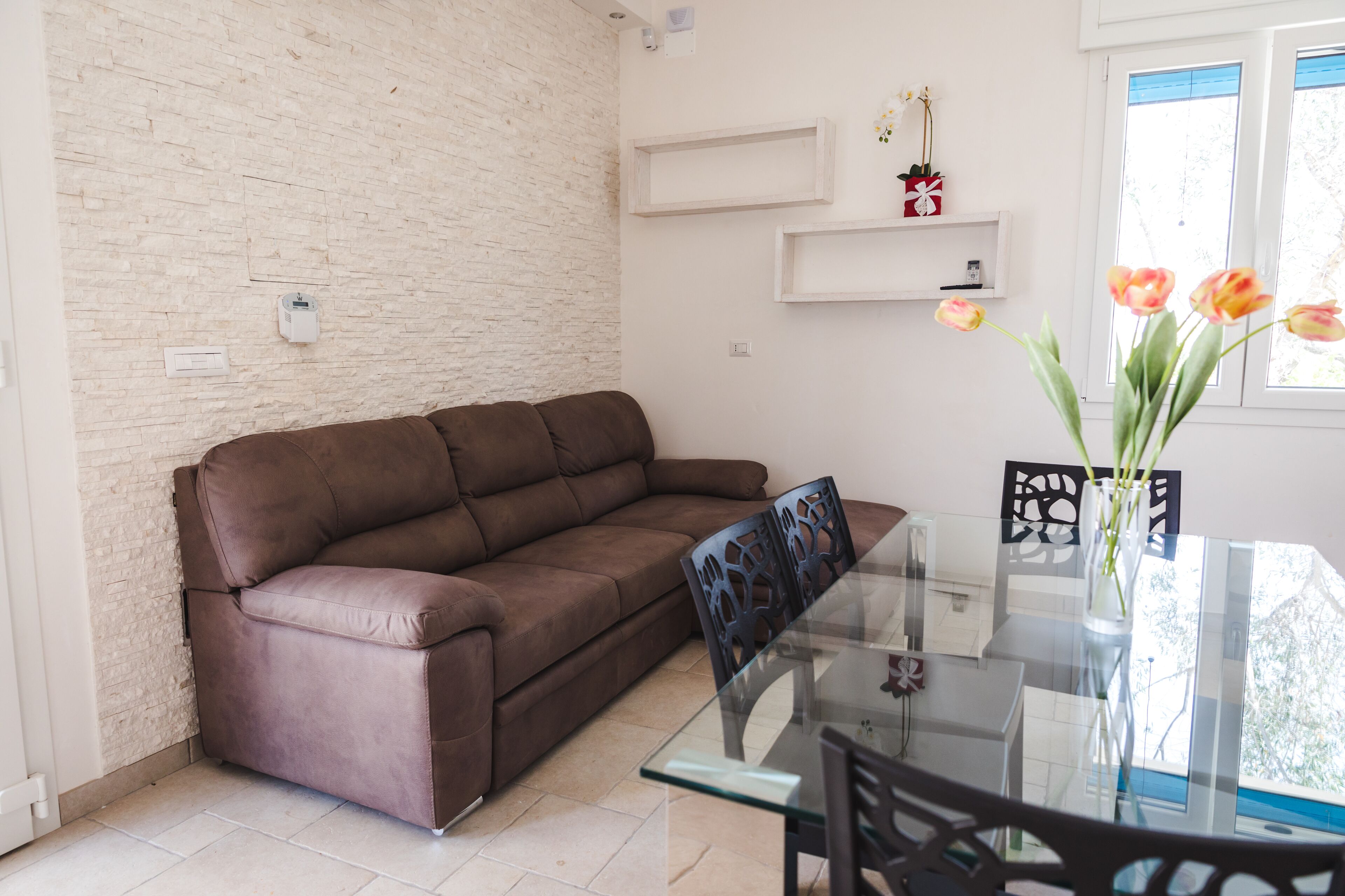 Un salón con un sofá marrón, una mesa de comedor de vidrio y sillas. Hay estantes blancos con objetos decorativos y una ventana con vistas.