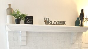 A white brick fireplace with a black glass door, a welcome sign above it, and a potted plant on the mantel.